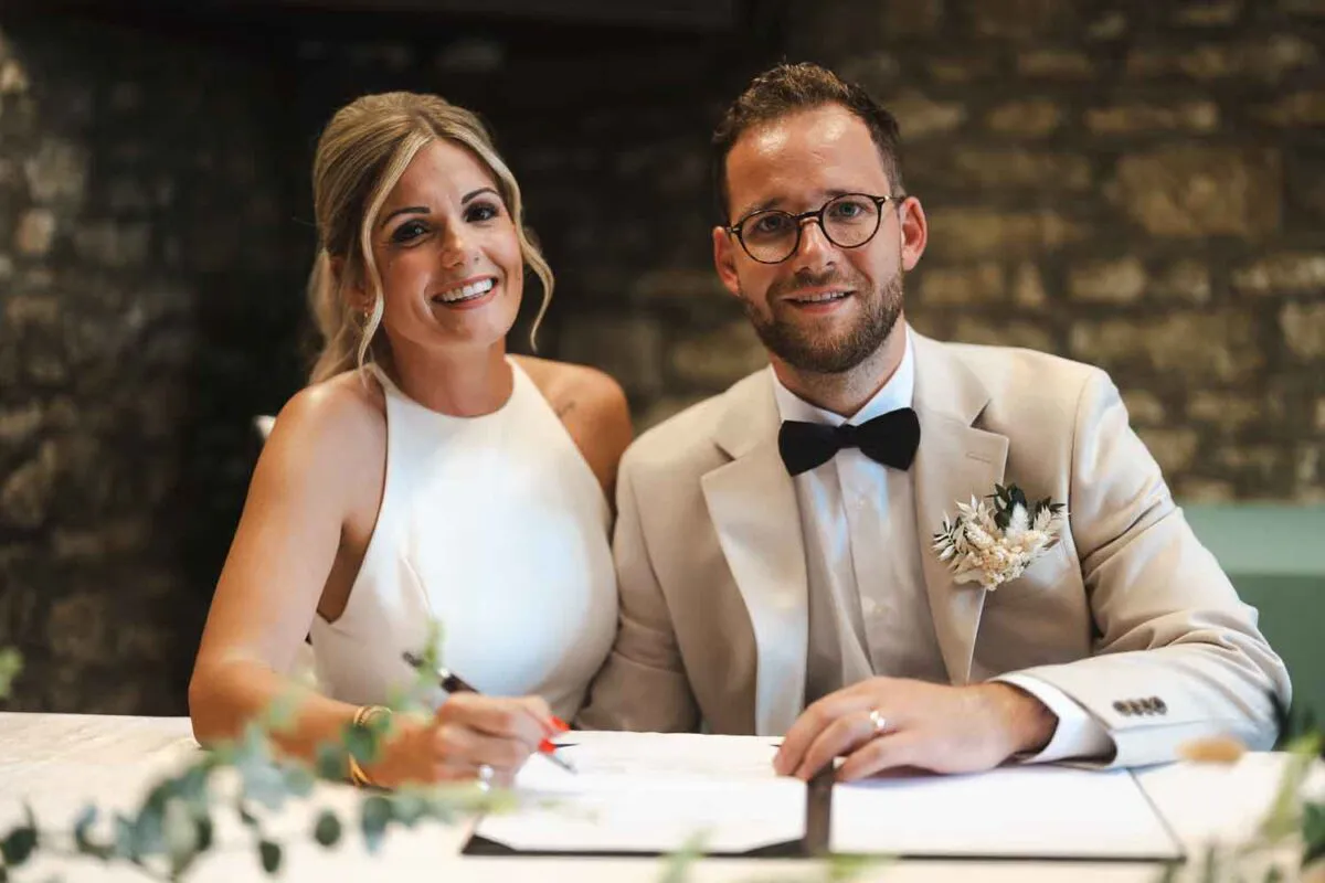A smiling bride in a halterneck dress and a groom in a beige suit with a black bow tie signing the wedding register together in front of an exposed stone wall.