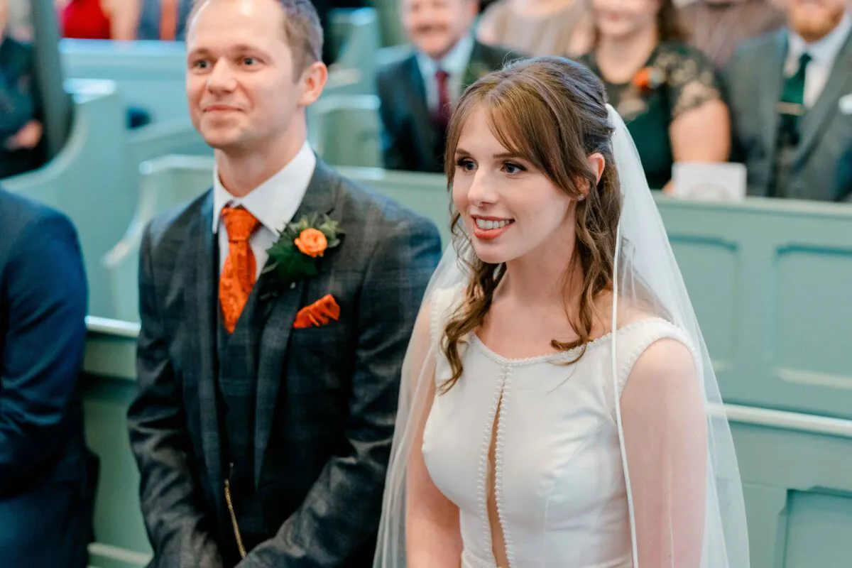 A smiling bride and groom sitting together in a church pews during their wedding ceremony, with the bride wearing a white gown and veil and the groom in a dark suit with an orange tie.