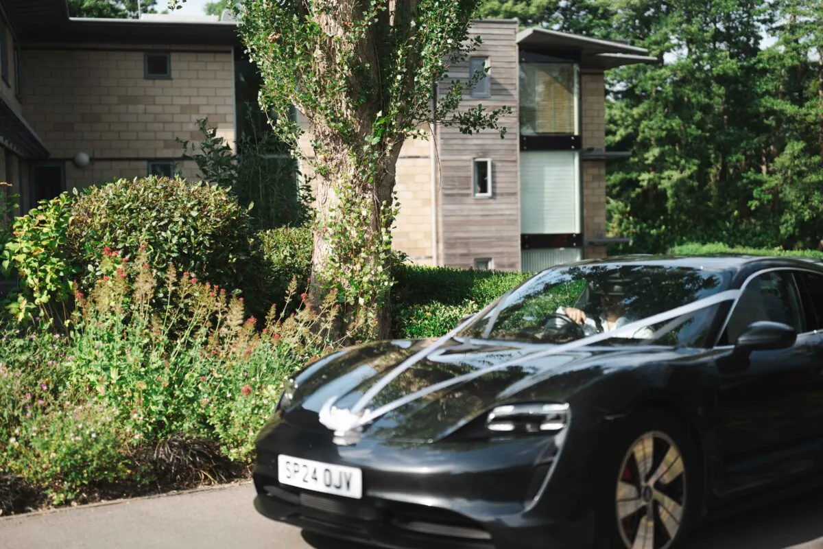 A sleek black wedding car decorated with white ribbons arrives at a modern hotel venue on a bright summer day.