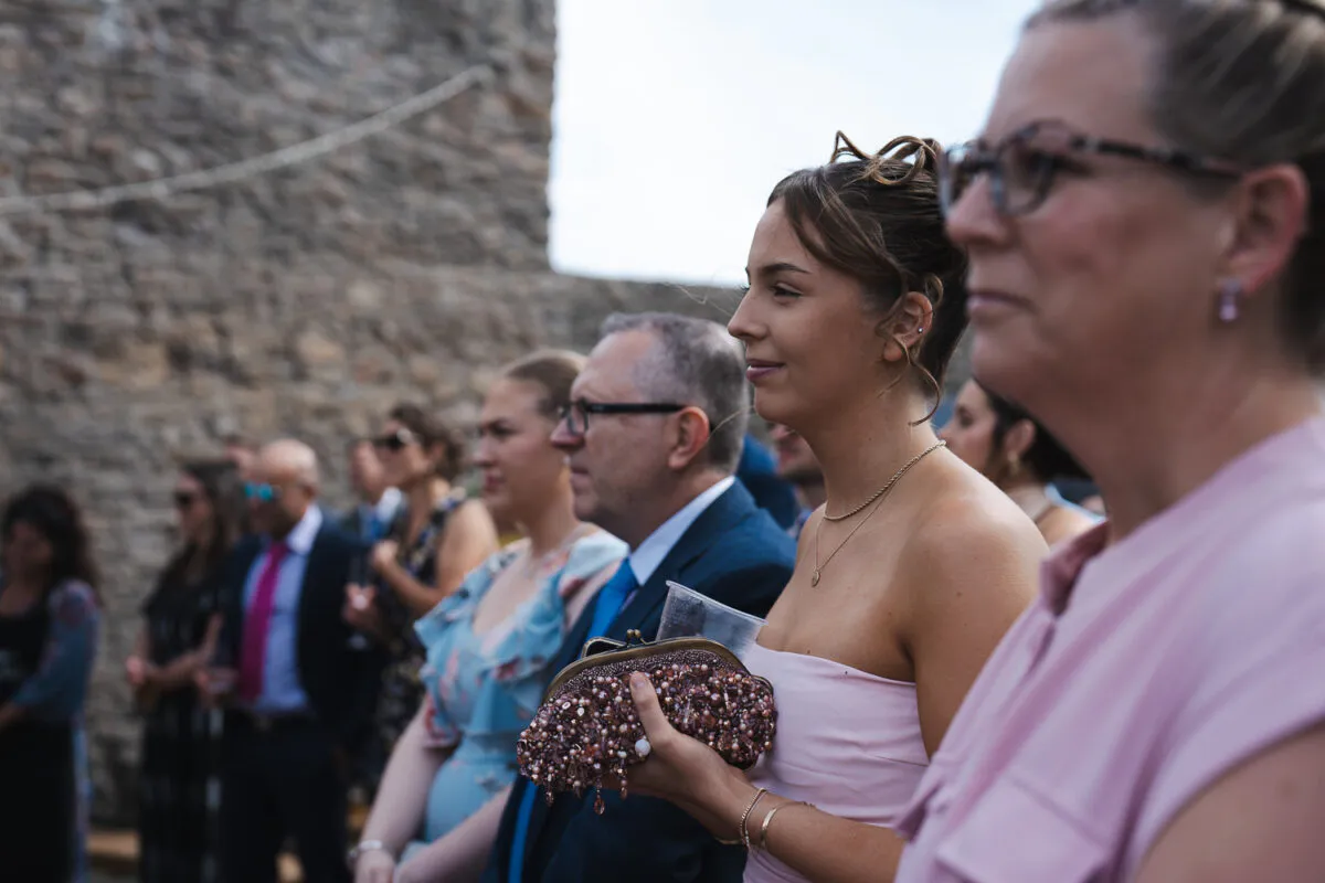 Wedding guests stand outside in profile, with a woman in a pink strapless top holding a beaded clutch in the foreground and a stone wall in the background.