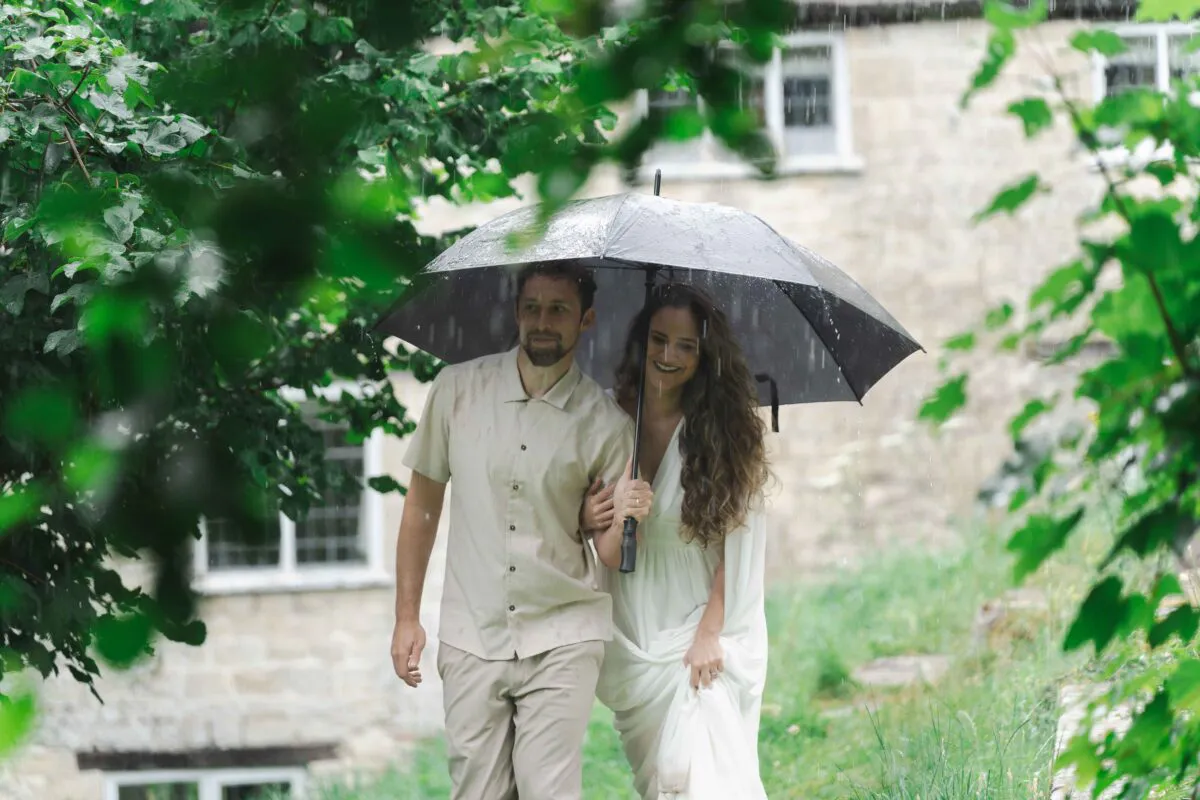 A newlywed couple smiling while walking together beneath a large umbrella during a gentle rain shower near a rustic stone building framed by green leaves.