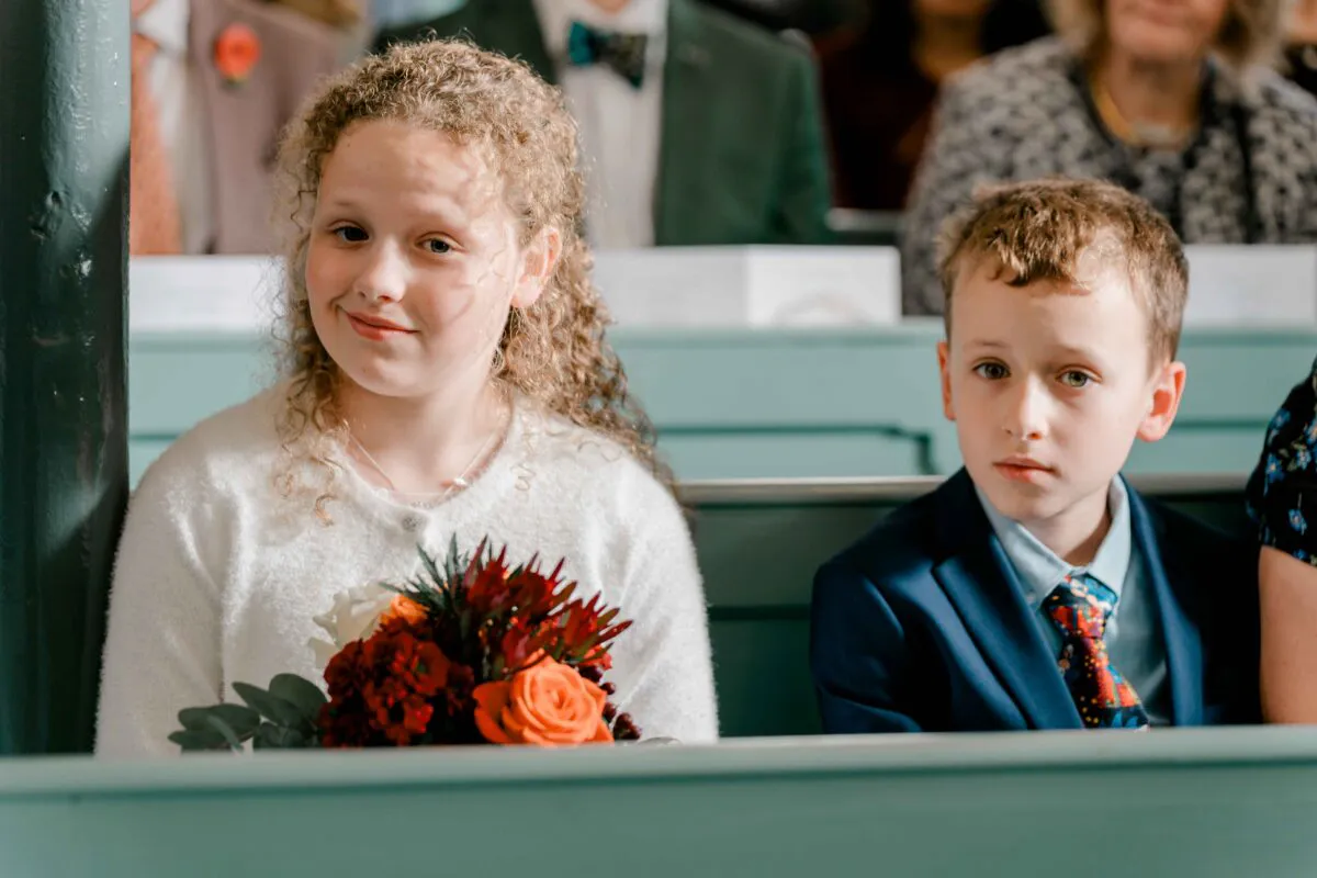 A young flower girl and page boy sit attentively on wooden pews during a wedding ceremony, holding a vibrant floral bouquet.