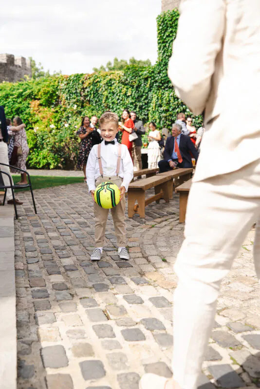 A young page boy in a white shirt and braces smiling happily while holding a football on a cobbled courtyard during an outdoor wedding reception.