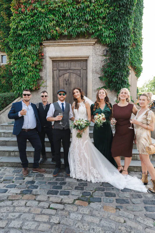 A newlywed couple and their wedding party pose together outside a historic stone building covered in lush green ivy.