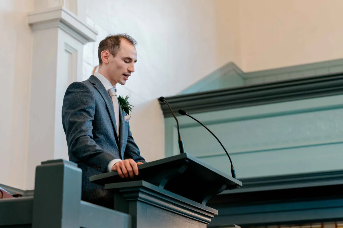 A groom delivering a speech from a pulpit inside a traditional church setting, wearing a tailored grey suit with a boutonniere.