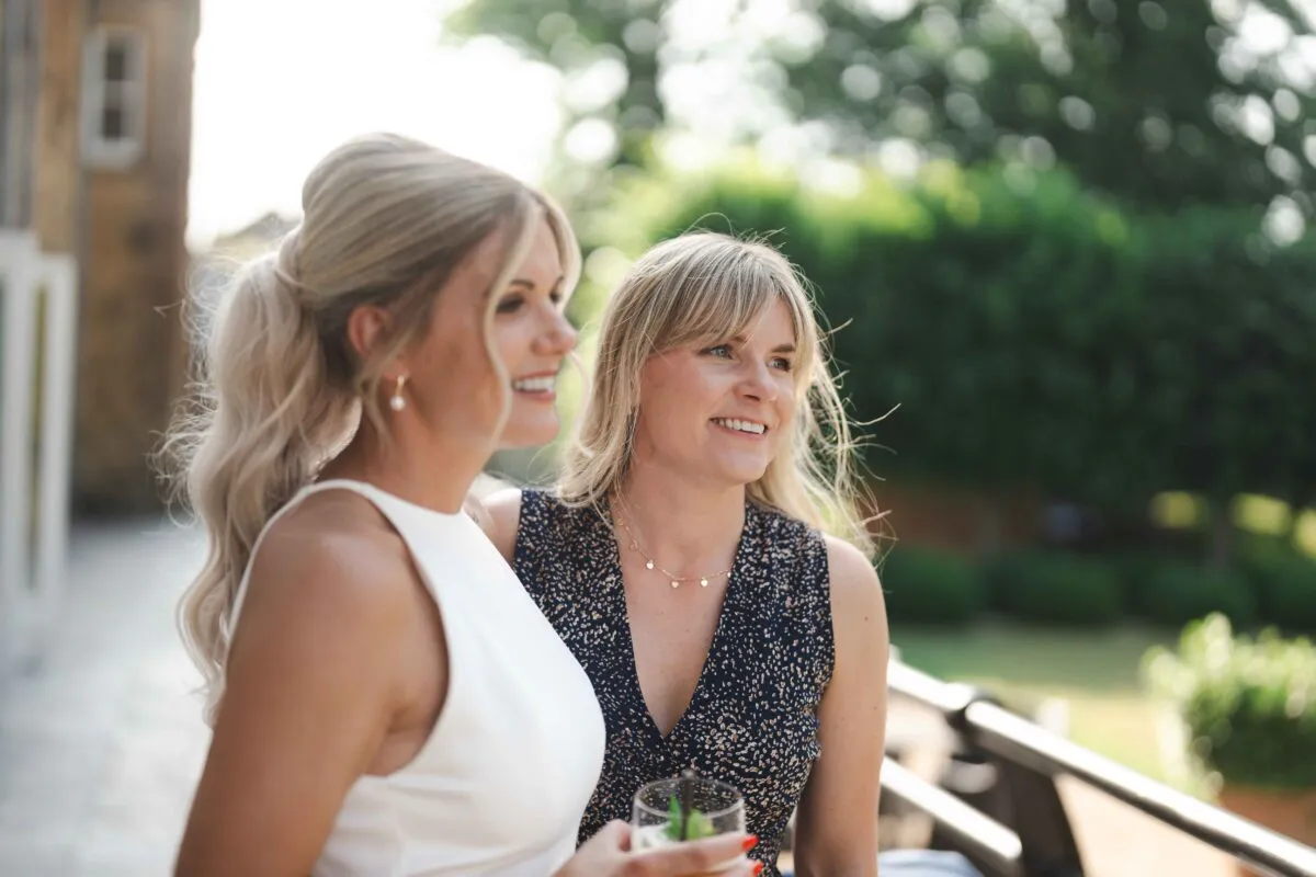 Two smiling wedding guests enjoy a drink together during the outdoor drinks reception on a bright sunny afternoon.