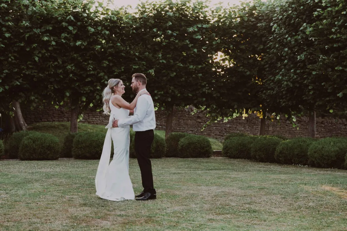 A newlywed couple laughing together during an outdoor golden hour portrait session against a backdrop of manicured topiary trees and a stone wall.