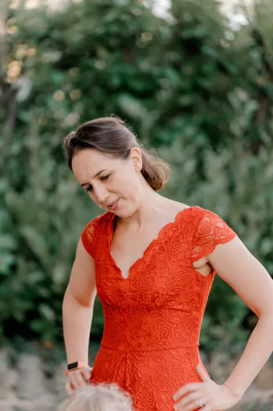 A wedding guest wearing a vibrant red lace dress with cap sleeves stands outdoors with a soft-focus green foliage background.