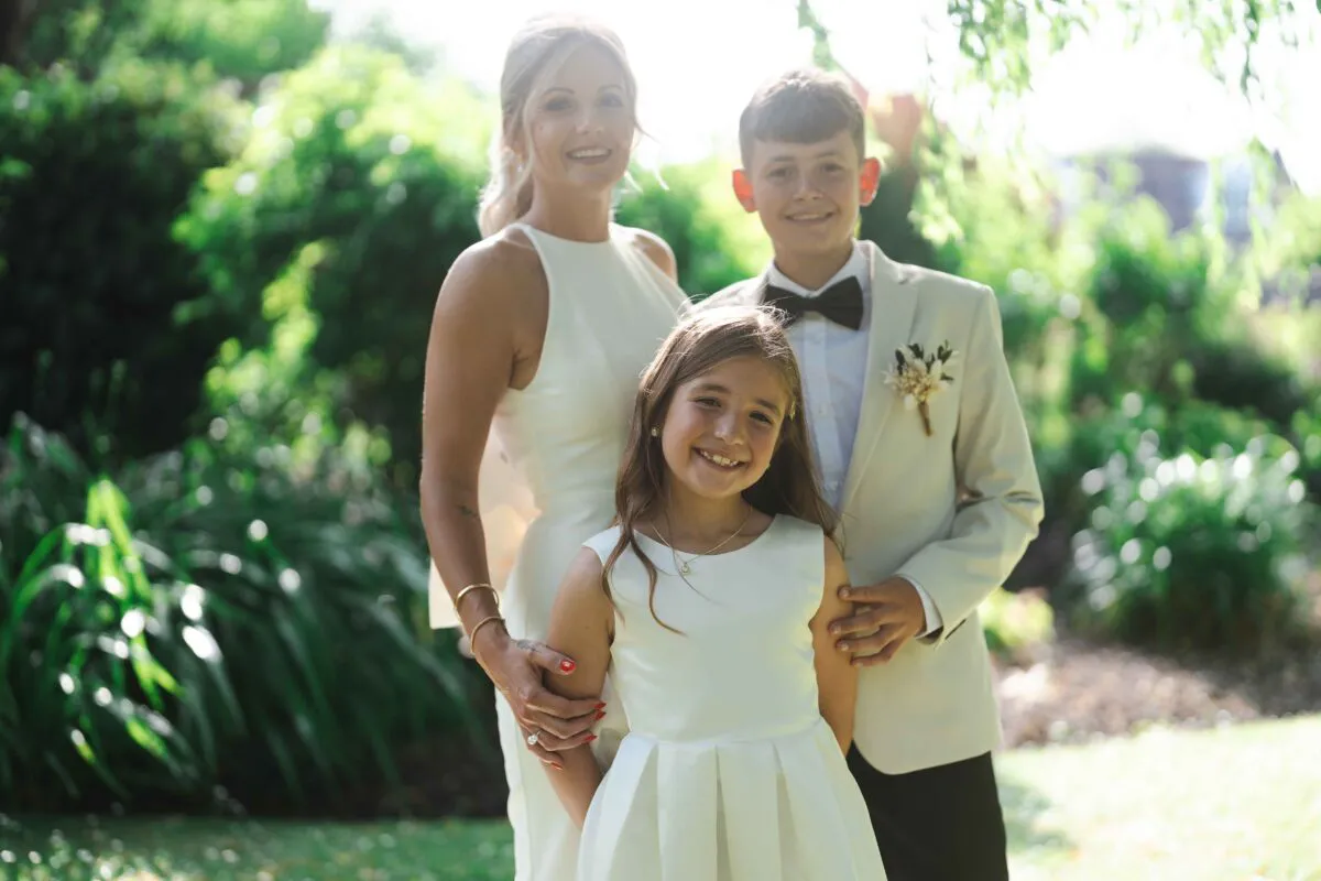 A smiling bride posing outdoors in a lush garden alongside a young flower girl in a white dress and a page boy in a beige suit.