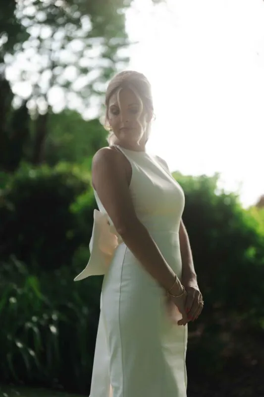 A beautiful backlit portrait of a bride outdoors in a garden setting, wearing a sleek halter neck column dress with an oversized bow detail.
