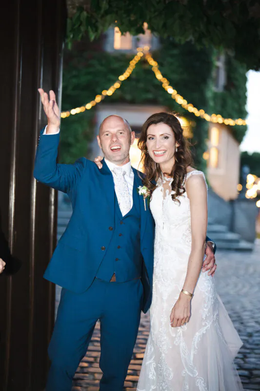 A joyful newlywed couple smiling and waving outdoors at dusk, with the groom wearing a blue three-piece suit and the bride in a lace sheath dress.