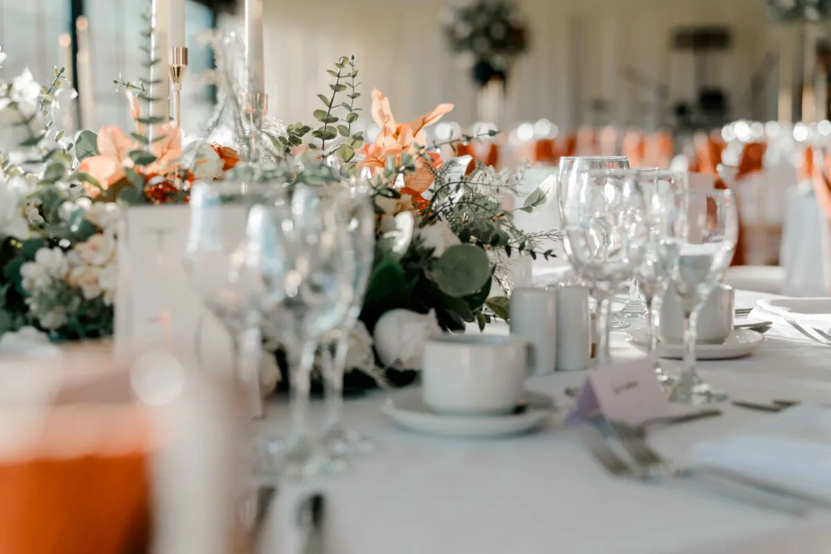A beautifully styled wedding reception tablescape features crisp white linens, delicate crystal glassware, and an abundant floral centrepiece with orange blooms and eucalyptus foliage.