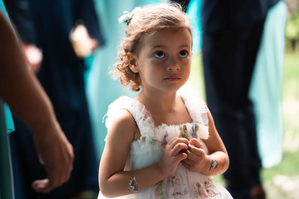 A young flower girl in a ruffled floral dress looks up curiously during the wedding ceremony.