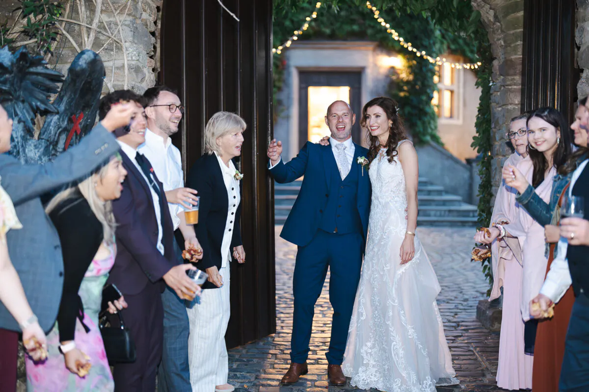 A smiling bride in a lace gown and a groom in a blue suit walk through a stone doorway into a cobbled courtyard while guests cheer.