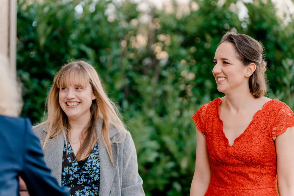 Two smiling wedding guests engage in conversation outdoors in front of a green leafy backdrop.