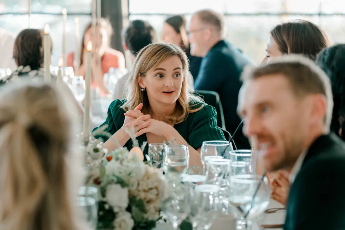A guest in a green dress sits at a banquet table engaged in conversation during a wedding breakfast.