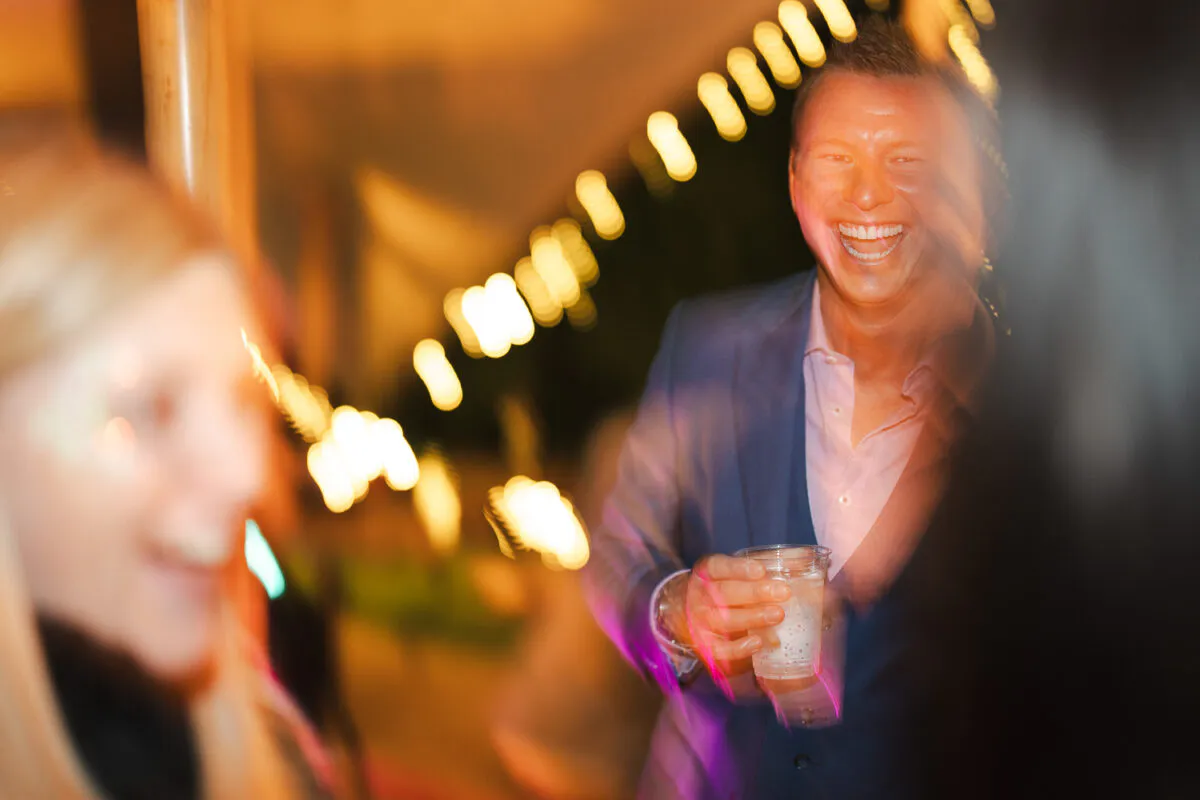 A guest in a navy suit and open-collared shirt laughs heartily while holding a drink at a lively evening wedding party illuminated by string lights.