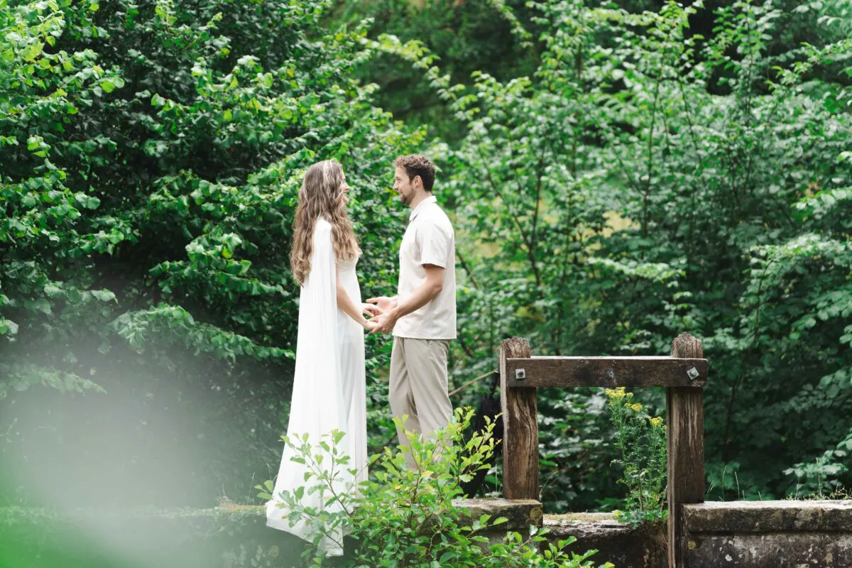 A newlywed couple holding hands and looking at each other on a stone and wooden bridge surrounded by lush green woodland.