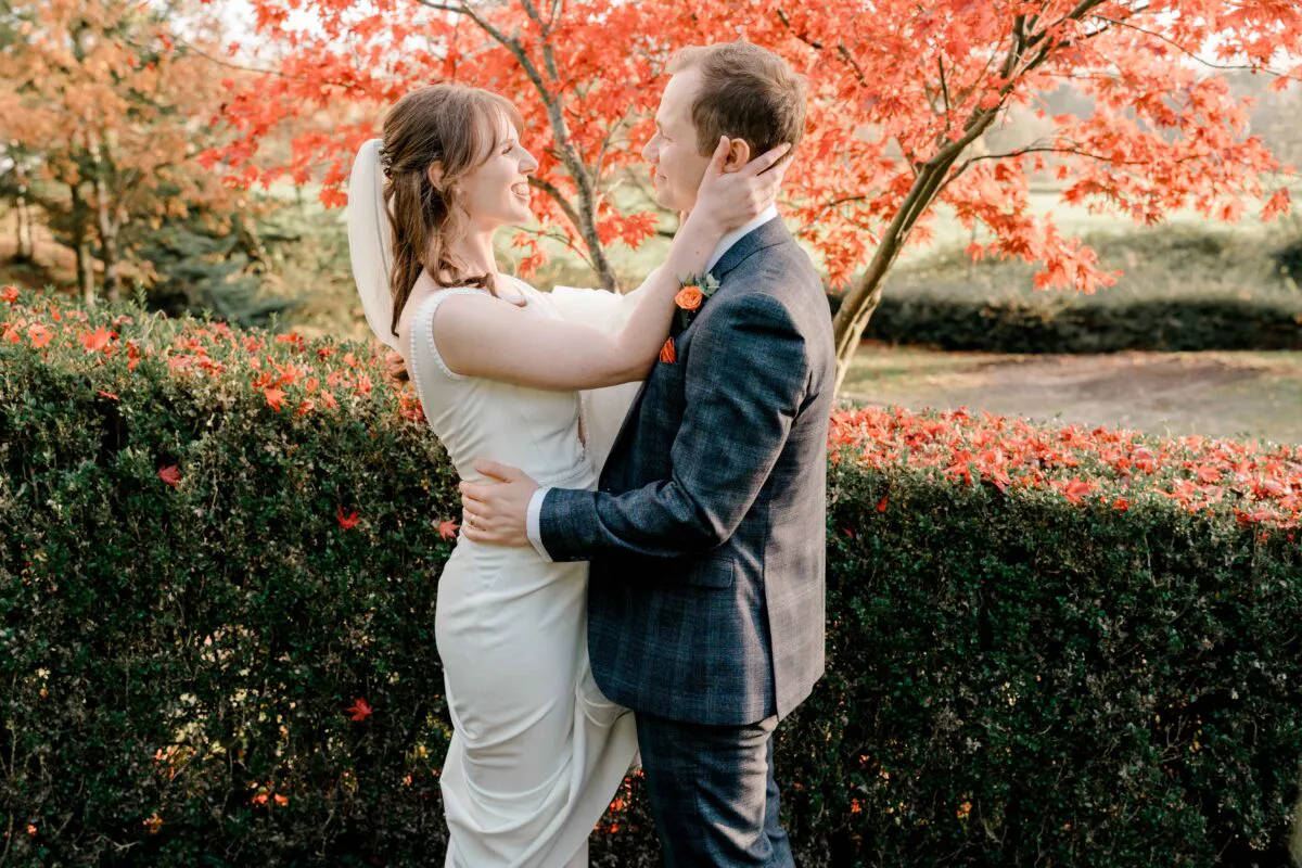 A newlywed couple share a tender moment facing each other in an outdoor garden setting surrounded by vibrant autumn foliage.