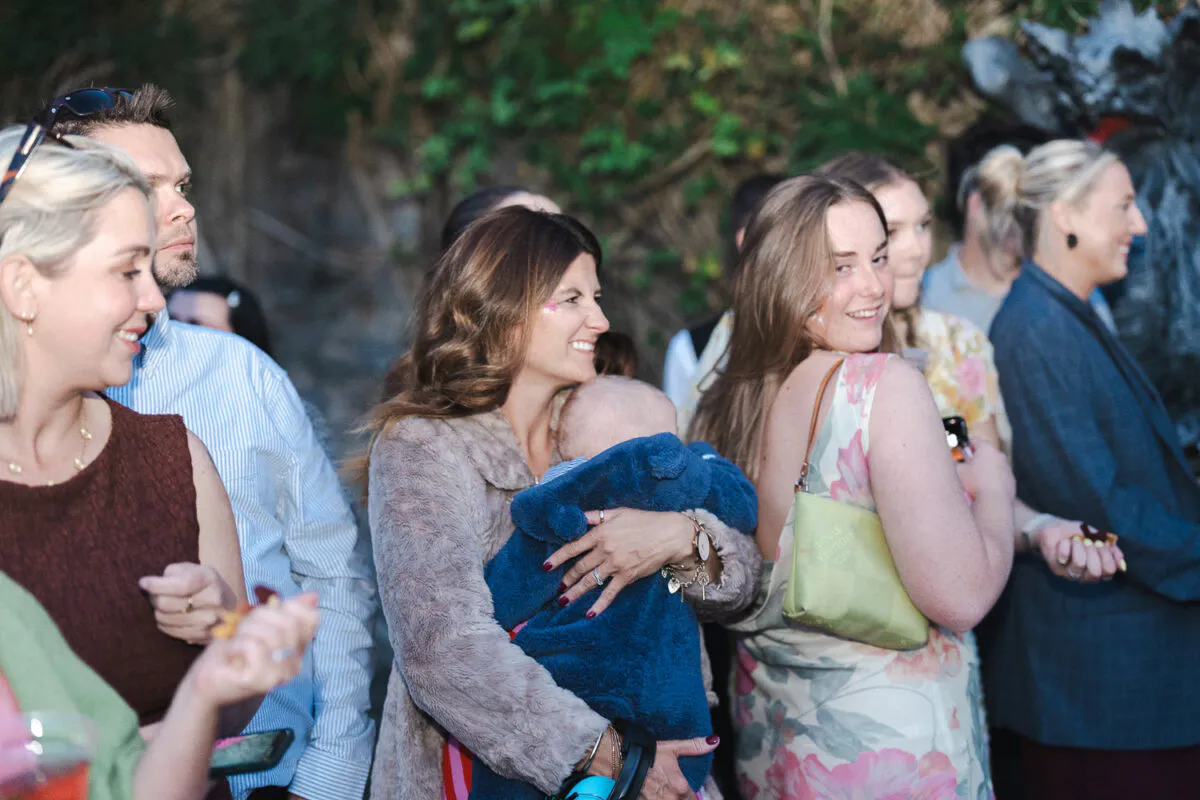 A candid outdoor photograph of wedding guests gathered together, including a woman holding a baby wrapped in a blue blanket.