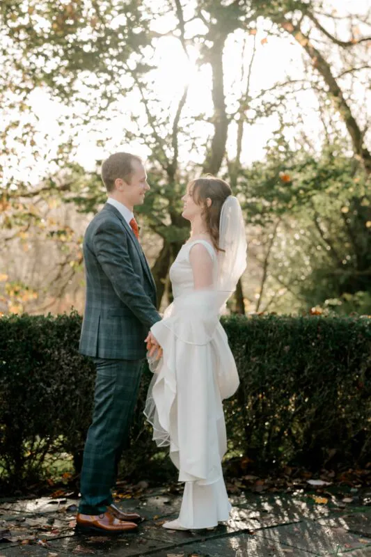A newlywed couple stands outdoors holding hands in front of a manicured hedge with sunlight filtering through autumn trees behind them.