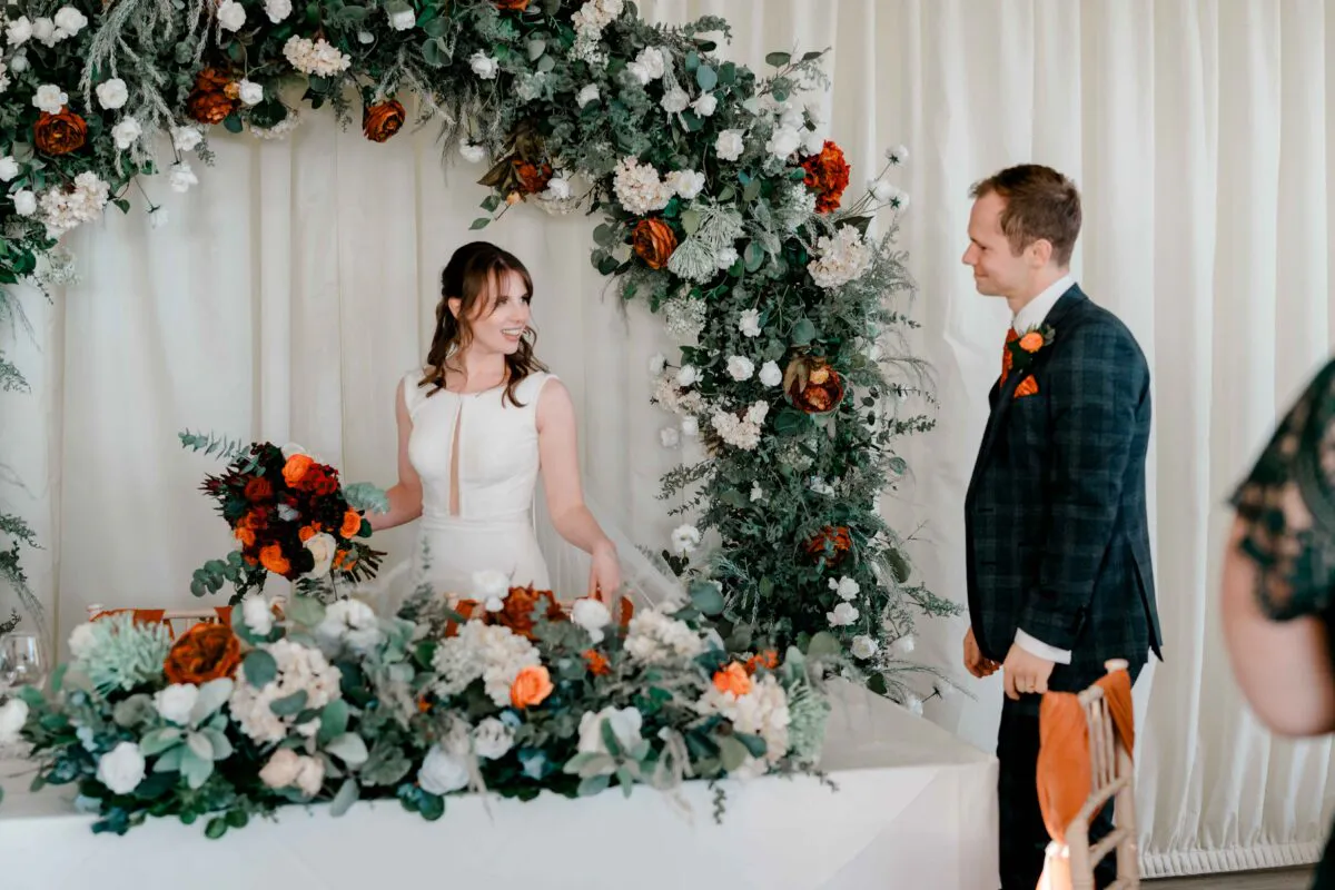 A smiling bride holding a vibrant bouquet stands near a lavish floral arch while looking towards the groom in his checked suit by the top table.