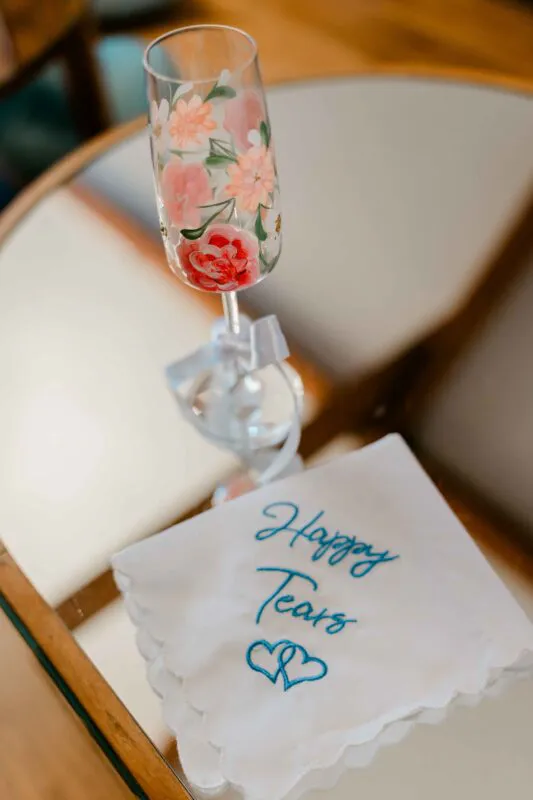 An intricately hand-painted floral champagne flute sits next to a white embroidered handkerchief reading happy tears with blue hearts.