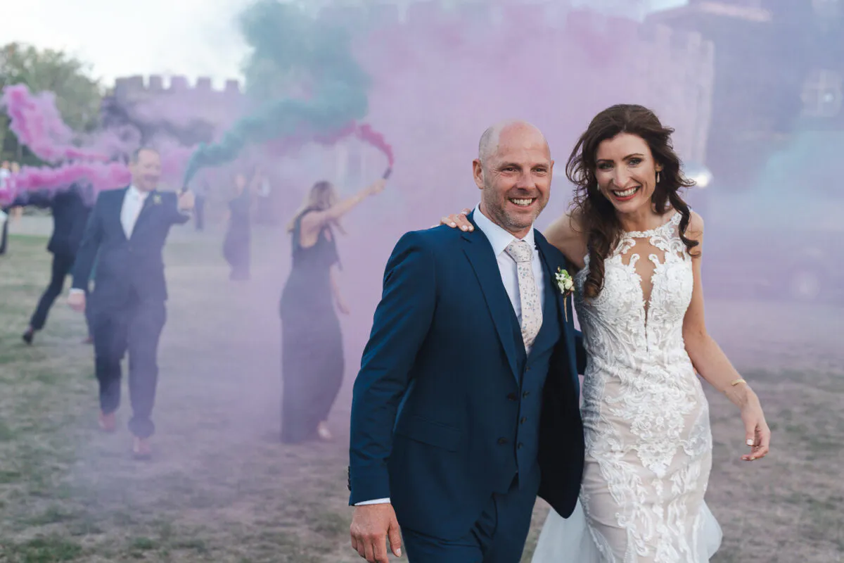 A smiling bride and groom walk outdoors in front of a castle while wedding guests set off pink and green smoke bombs behind them.