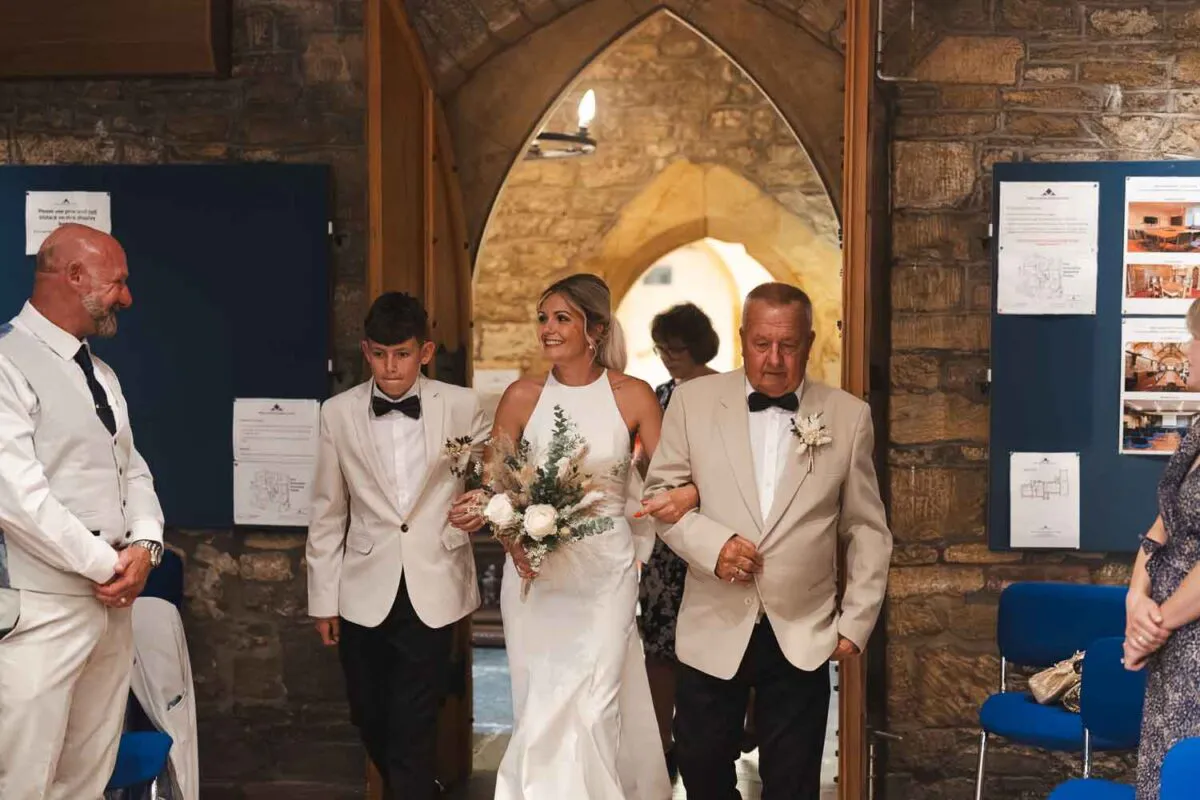 The radiant bride makes her grand entrance walking down the aisle inside a historic stone church, escorted by family members while holding a textured bouquet.