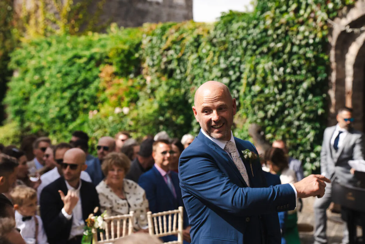 A smiling groom in a navy suit gestures towards guests seated outdoors in front of an ivy-covered stone wall during a wedding ceremony.