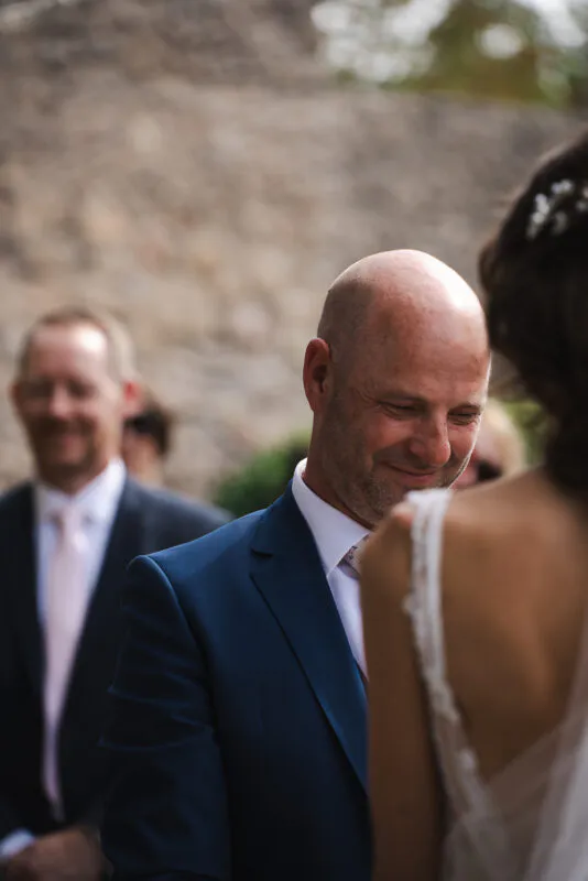 An emotional groom in a navy suit shares a tender moment with his bride, looking down with a gentle smile during their wedding ceremony.