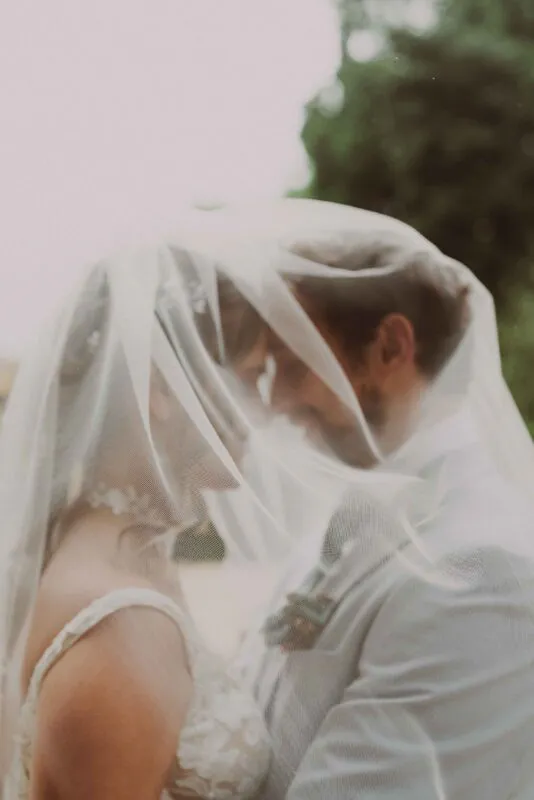 A newlywed couple share a tender moment standing close together underneath a sheer veil outdoors.