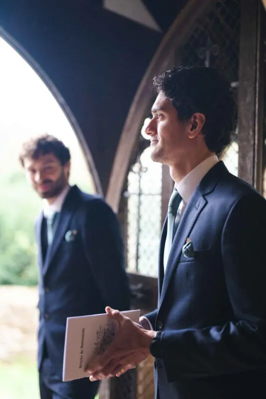 A groom in a sharp navy suit stands near an arched doorway holding a ceremony booklet, with another groomsman softly blurred in the background.