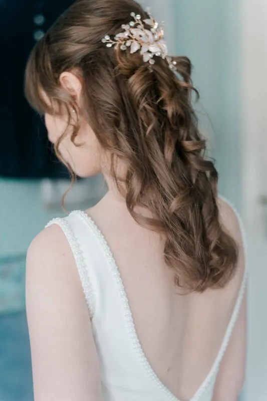 A close-up back view of a bride showcasing her half-up curly hairstyle adorned with a delicate gold and pearl hair comb, wearing a white gown with a beaded V-back neckline.