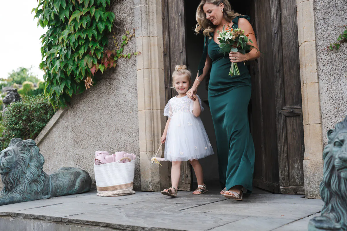 A bridesmaid in a green satin gown and a young flower girl walk hand in hand out of a stone doorway carrying a bouquet and a wand.