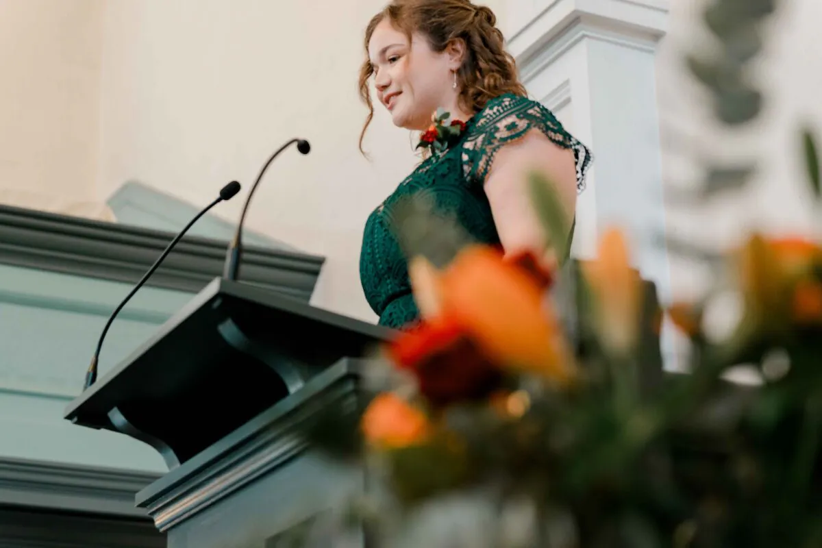 A smiling bride wearing an emerald green lace dress delivers a speech from a lectern adorned with microphones during the wedding reception.
