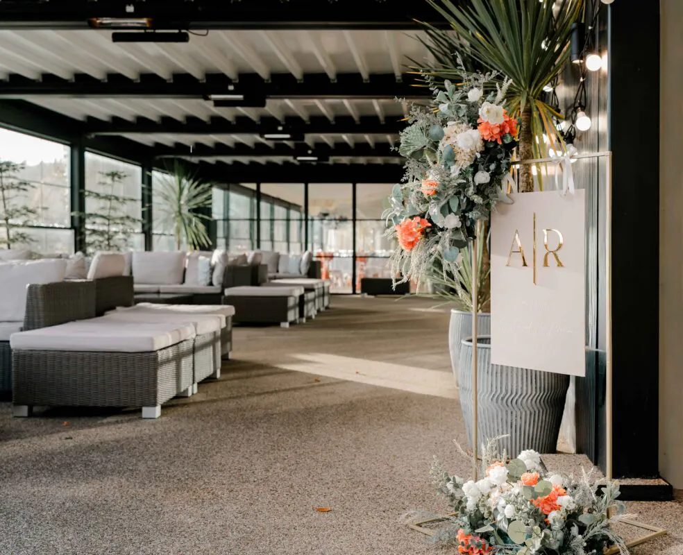 An elegant modern wedding welcome sign stands adorned with peach and white floral arrangements on a copper frame, set on a bright terrace with lounge seating.