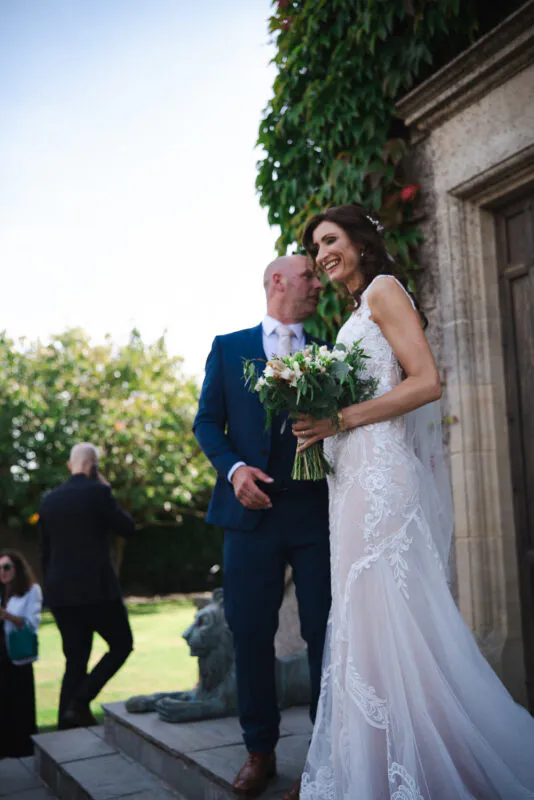 A laughing bride holding a hand-tied bouquet stands next to her groom in a navy suit outside a historic stone building.