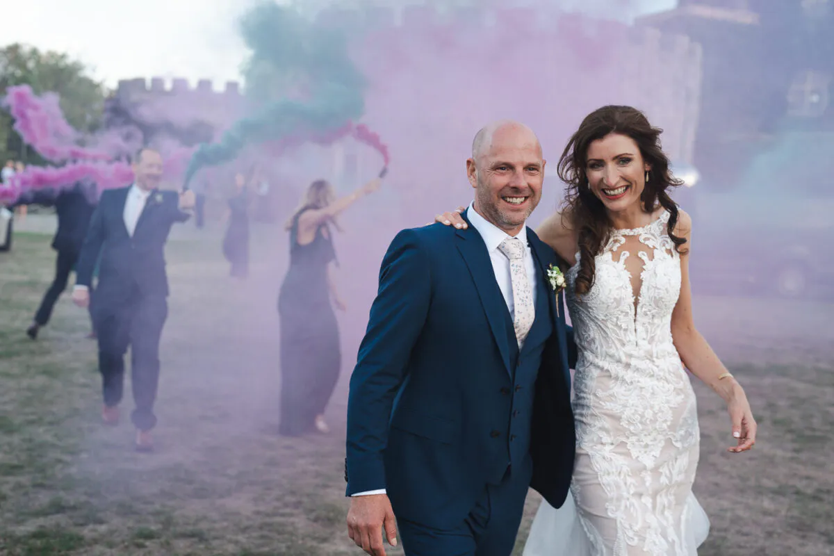 A smiling bride and groom walk outdoors against a castle backdrop while wedding guests set off colourful smoke bombs behind them.