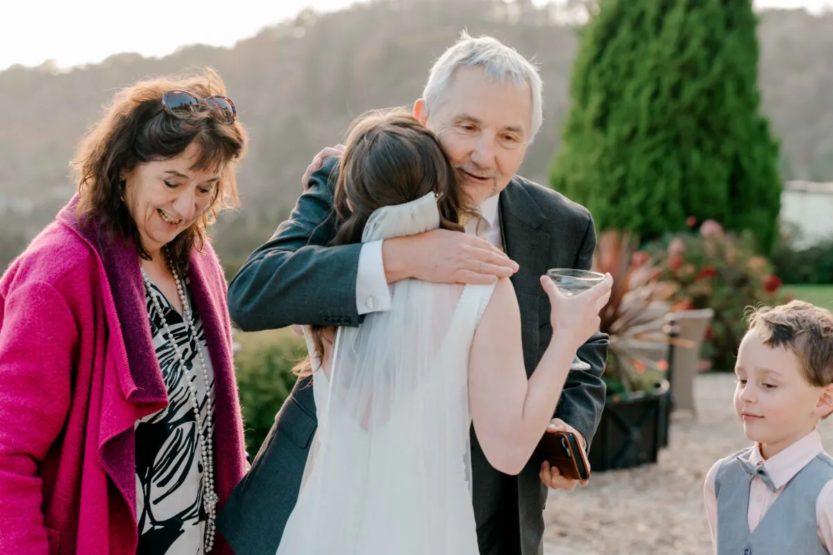 An emotional father warmly embraces the bride outdoors during the wedding reception while holding a champagne coupe, alongside family members including the mother of the bride.