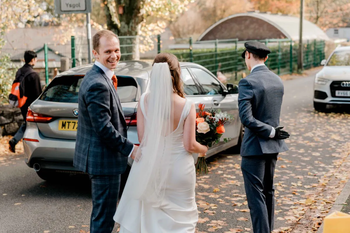 A smiling groom looks back over his shoulder while walking hand in hand with his bride towards a waiting wedding car on an autumn street covered in fallen leaves.