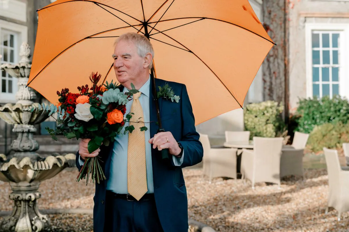 An older man in a navy suit and yellow tie stands outdoors holding a bright orange umbrella and a vibrant floral bouquet.