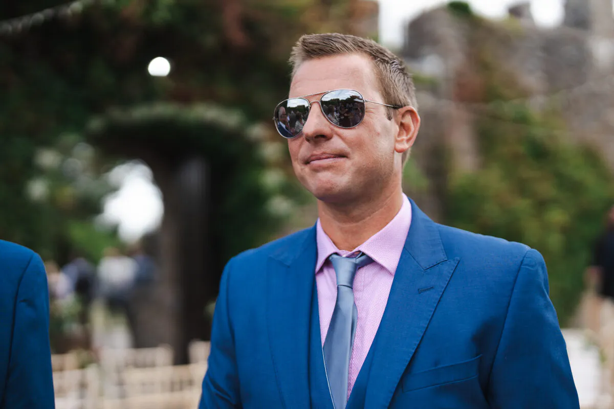 A groom wearing a blue suit, pink shirt, and sunglasses stands outdoors near a stone archway during a wedding ceremony.