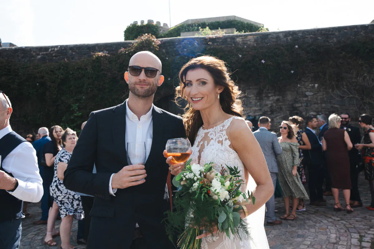 A smiling bride holding a textured bouquet and a groom in a dark suit with sunglasses stand together outdoors in a cobbled courtyard during their wedding drinks reception.