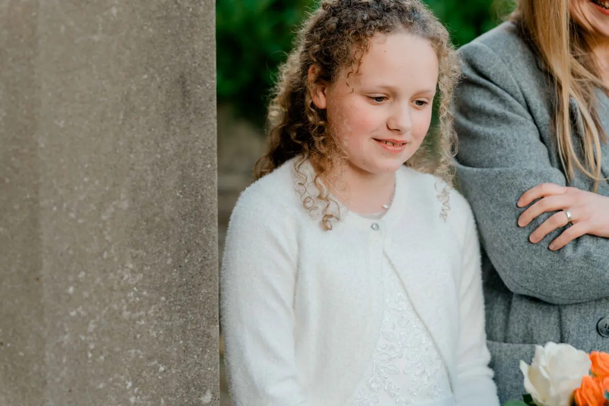 A young flower girl with curly hair, wearing a white cardigan and patterned dress, stands smiling next to a guest in a grey coat holding a bright floral bouquet.