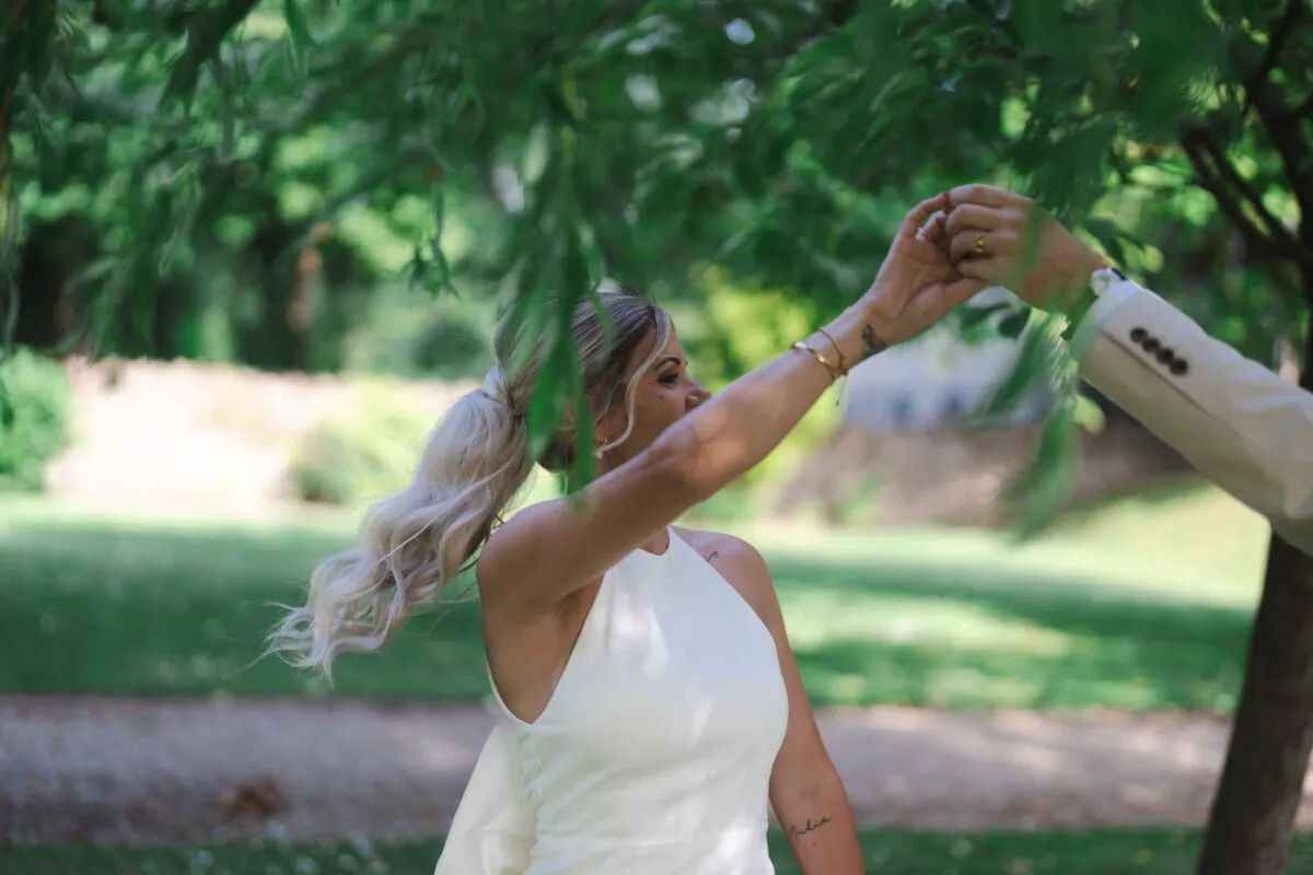 A bride in a sleek halterneck gown holds hands with the groom beneath the leafy canopy of an ancient tree on a sunny wedding day.