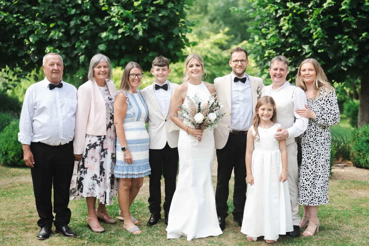A formal family group photograph featuring the bride and groom standing outdoors in a lush garden setting surrounded by relatives and a young flower girl.