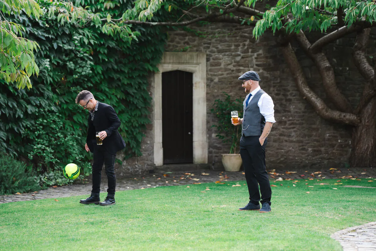 Two grooms in smart casual attire enjoy a drink together on a lawn outside a stone manor house while kicking a bright yellow ball.