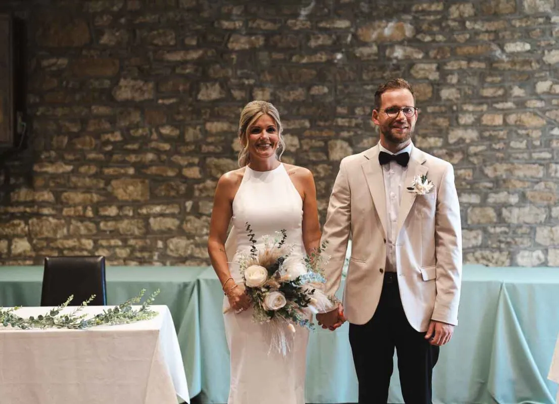 A newlywed couple holds hands and smiles in front of a rustic exposed stone wall inside a barn venue, with the bride holding a neutral bouquet featuring white roses and dried grasses.