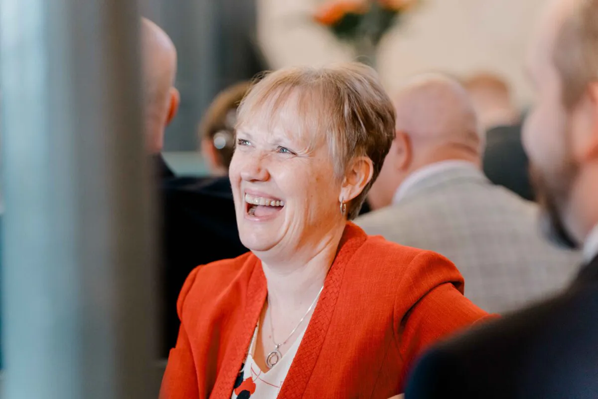 A mother of the bride laughs heartily during wedding speeches, wearing a bright orange blazer and silver pendant necklace in a candid indoor moment.